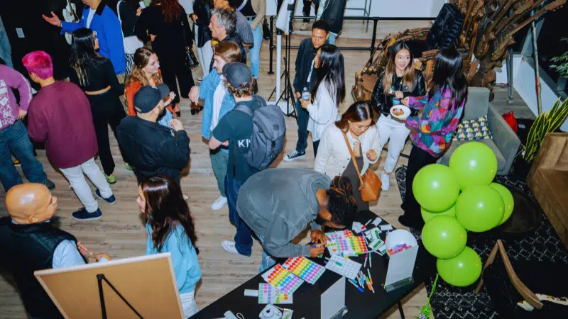 A group of people socializing at an indoor event, with some gathered around a table covered in colorful stickers and green balloons.