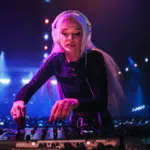 A DJ with long blonde hair and headphones mixing music on a controller at a nightclub, illuminated by colorful stage lights, turns every beat into a ritual of sound and energy.