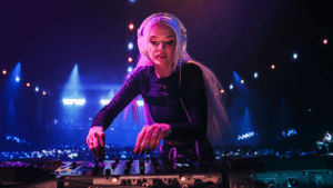 A DJ with long blonde hair and headphones mixing music on a controller at a nightclub, illuminated by colorful stage lights, turns every beat into a ritual of sound and energy.