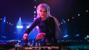 A DJ with long blonde hair and headphones mixing music on a controller at a nightclub, illuminated by colorful stage lights, turns every beat into a ritual of sound and energy.