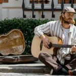 A man wearing a cap sits on a sidewalk, playing an acoustic guitar next to an open guitar case—inviting passersby to support his music through spontaneous crowdfunding.