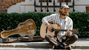 How to Use Crowdfunding to Launch Your Next Project A man wearing a cap sits on a sidewalk, playing an acoustic guitar next to an open guitar case—inviting passersby to support his music through spontaneous crowdfunding.