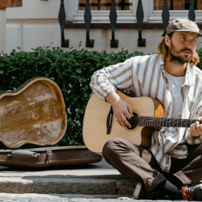 A man wearing a cap sits on a sidewalk, playing an acoustic guitar next to an open guitar case—inviting passersby to support his music through spontaneous crowdfunding.