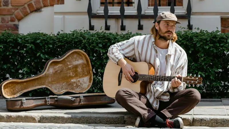 A man wearing a cap sits on a sidewalk, playing an acoustic guitar next to an open guitar case—inviting passersby to support his music through spontaneous crowdfunding.