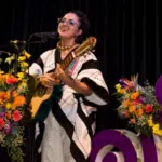 A woman wearing a traditional LATAM outfit plays a guitar and sings into a microphone on stage, surrounded by colorful floral arrangements and purple decorations.