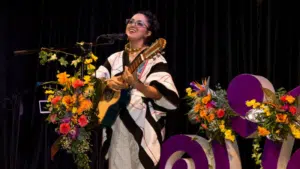 A woman wearing a traditional LATAM outfit plays a guitar and sings into a microphone on stage, surrounded by colorful floral arrangements and purple decorations.