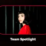 A person with a short dark bob haircut and bangs, wearing a red top, stands in front of vertical black bars. The text reads, "Team Spotlight.
