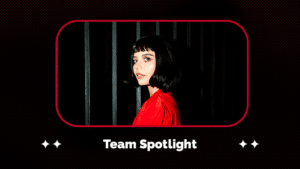 A person with a short dark bob haircut and bangs, wearing a red top, stands in front of vertical black bars. The text reads, "Team Spotlight.