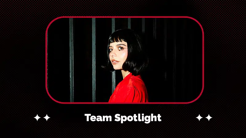 A person with a short dark bob haircut and bangs, wearing a red top, stands in front of vertical black bars. The text reads, "Team Spotlight.