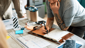 A person writes in a notebook on a desk with a calendar, documents, labels, water bottle, and stationery during a meeting or planning session.