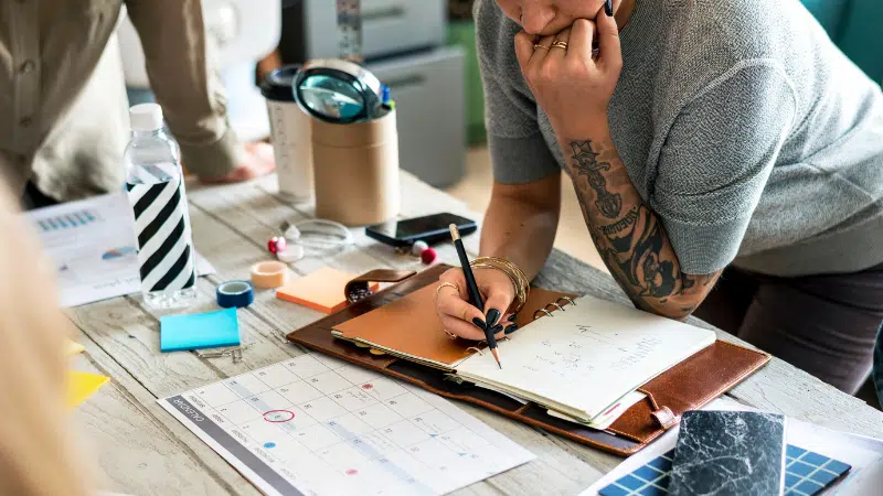 A person writes in a notebook on a desk with a calendar, documents, labels, water bottle, and stationery during a meeting or planning session.