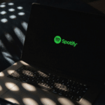 A laptop displaying the green Spotify logo on its screen sits on a bed with dappled sunlight and shadow patterns, hinting at a cozy space where music and AI technology meet.