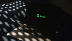 Spotify’s New AI Protections for Artists: Reinforcing Our Commitment to Transparency A laptop displaying the green Spotify logo on its screen sits on a bed with dappled sunlight and shadow patterns, hinting at a cozy space where music and AI technology meet.