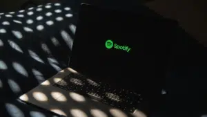 A laptop displaying the green Spotify logo on its screen sits on a bed with dappled sunlight and shadow patterns, hinting at a cozy space where music and AI technology meet.
