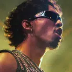A person wearing sunglasses and a chain-adorned top performs onstage under dramatic lighting.