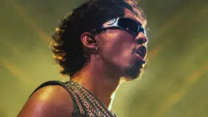 A person wearing sunglasses and a chain-adorned top performs onstage under dramatic lighting.