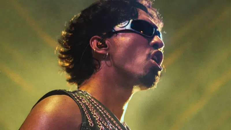 A person wearing sunglasses and a chain-adorned top performs onstage under dramatic lighting.