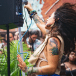 A singer with curly hair and tattoos performs energetically outdoors in front of a crowd, holding a microphone with one hand raised. The good vibes are contagious as people in the background watch and smile.