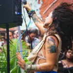 A singer with curly hair and tattoos performs energetically outdoors in front of a crowd, holding a microphone with one hand raised. The good vibes are contagious as people in the background watch and smile.
