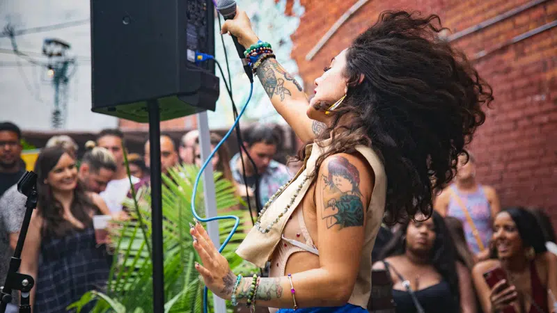 A singer with curly hair and tattoos performs energetically outdoors in front of a crowd, holding a microphone with one hand raised. The good vibes are contagious as people in the background watch and smile.
