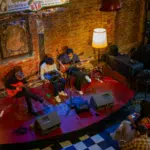 Three musicians play guitars and keyboard on a small stage in a cozy, dimly lit Bogotá venue with red brick walls and vintage decor.