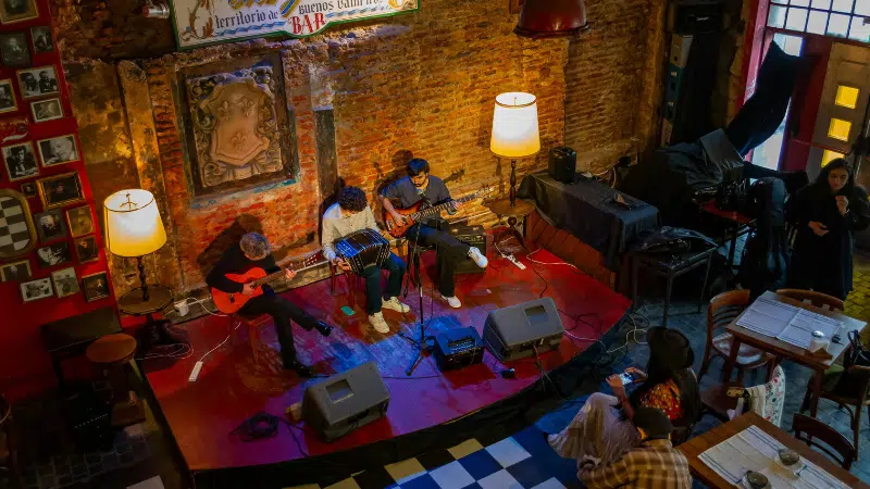 Three musicians play guitars and keyboard on a small stage in a cozy, dimly lit Bogotá venue with red brick walls and vintage decor.