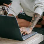 A person with tattoos is sitting cross-legged on a couch using digital tools—a laptop on the table—while another person in the background holds a tablet.