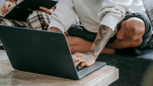 A person with tattoos is sitting cross-legged on a couch using digital tools—a laptop on the table—while another person in the background holds a tablet.