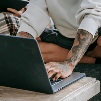 A person with tattoos is sitting cross-legged on a couch using digital tools—a laptop on the table—while another person in the background holds a tablet.