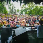 A DJ performs on an outdoor stage to a large crowd of people during the day, surrounded by trees and greenery, capturing the vibrant energy of festivals.