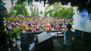 A DJ performs on an outdoor stage to a large crowd of people during the day, surrounded by trees and greenery, capturing the vibrant energy of festivals.