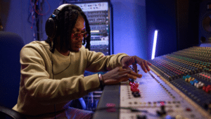 A person wearing headphones and sunglasses adjusts controls on a large audio mixing console, expertly managing audio content in a professional recording studio.