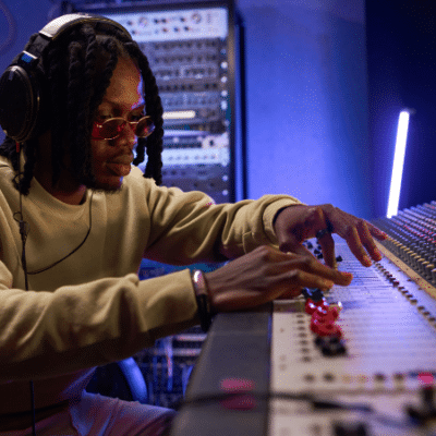 A person wearing headphones and sunglasses adjusts controls on a large audio mixing console, expertly managing audio content in a professional recording studio.
