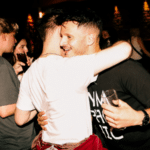 Two men hug and smile while holding drinks at a dimly lit social gathering in Dublin; other people are talking in the background.
