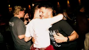 Two men hug and smile while holding drinks at a dimly lit social gathering in Dublin; other people are talking in the background.