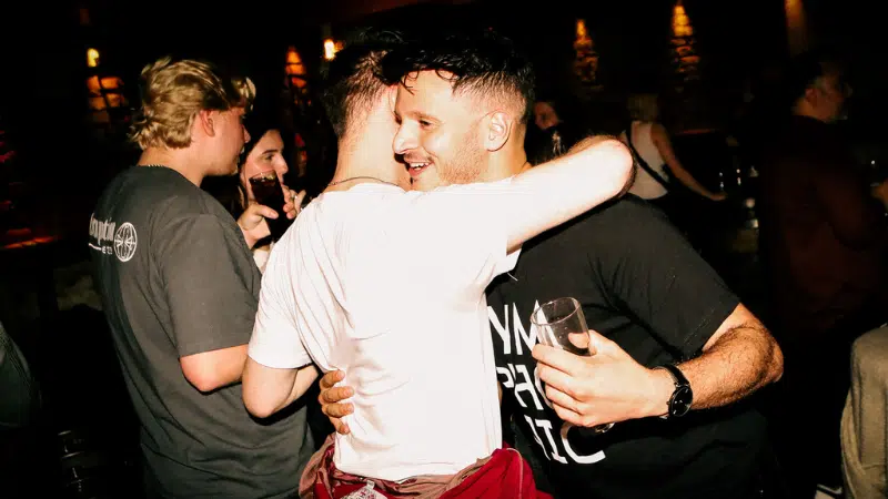 Two men hug and smile while holding drinks at a dimly lit social gathering in Dublin; other people are talking in the background.