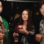 Three people stand together at a social event indoors, holding drinks and smiling at the camera. The background is dimly lit with other attendees visible.