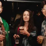 Three people stand together at a social event indoors, holding drinks and smiling at the camera. The background is dimly lit with other attendees visible.