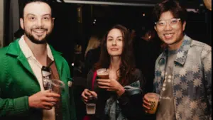 Three people stand together at a social event indoors, holding drinks and smiling at the camera. The background is dimly lit with other attendees visible.