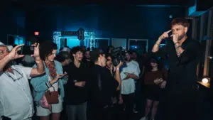 A man performs on stage to a crowd at a dimly lit indoor event; several people in the audience are taking photos and videos.