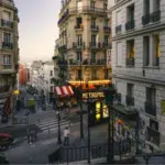 A street scene in Paris at dusk shows classic French architecture, a hotel, the MaMA Café Métropol sign, stairs, and people walking along the sidewalks.