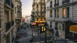 A street scene in Paris at dusk shows classic French architecture, a hotel, the MaMA Café Métropol sign, stairs, and people walking along the sidewalks.