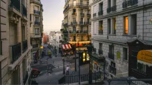 A street scene in Paris at dusk shows classic French architecture, a hotel, the MaMA Café Métropol sign, stairs, and people walking along the sidewalks.