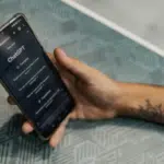 A person holding a smartphone displaying the ChatGPT website, with a geometric patterned table beneath their hand, showcases how ChatGPT integrates seamlessly into everyday life.