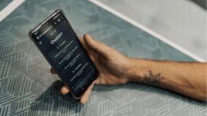 A person holding a smartphone displaying the ChatGPT website, with a geometric patterned table beneath their hand, showcases how ChatGPT integrates seamlessly into everyday life.