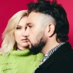 A woman with blond hair and a green sweater stands beside a man with short dark hair and a beard, against a solid magenta background.