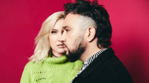 A woman with blond hair and a green sweater stands beside a man with short dark hair and a beard, against a solid magenta background.