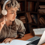 Introducing ‘Enrollments’ in the SymphonicMS A person with headphones studies at a desk, immersed in symphonic tunes, surrounded by a laptop, notebook, open book, and coffee cup in a quiet library or study room.