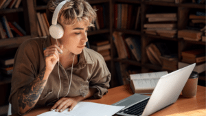 Introducing ‘Enrollments’ in the SymphonicMS A person with headphones studies at a desk, immersed in symphonic tunes, surrounded by a laptop, notebook, open book, and coffee cup in a quiet library or study room.