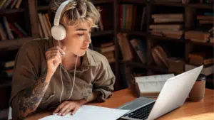 A person with headphones studies at a desk, immersed in symphonic tunes, surrounded by a laptop, notebook, open book, and coffee cup in a quiet library or study room.